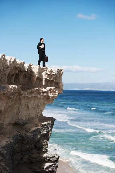 世界を征服できる 海を見下ろす崖の端に立っている本とブリーフケースを持っている若いビジネスマン — ストック写真