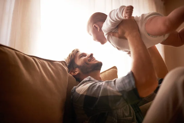 stock image Smile, father and lifting baby on sofa in home living room, playing or bonding together. Funny, care and dad holding infant, newborn or child on couch in lounge, having fun and enjoying quality time