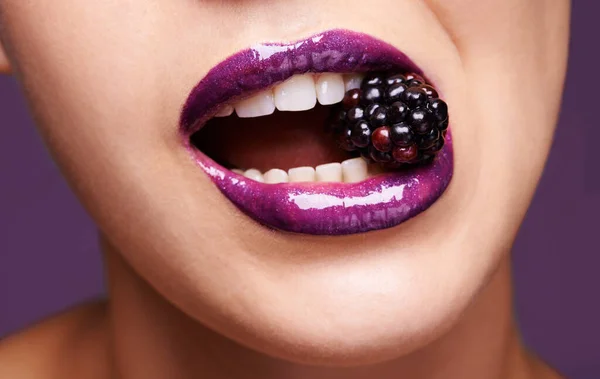 Woman lips, purple lipstick with blackberry and makeup, shine and creativity with beauty isolated on studio background. Closeup of fruit between female model teeth, cosmetic product and cosmetology.