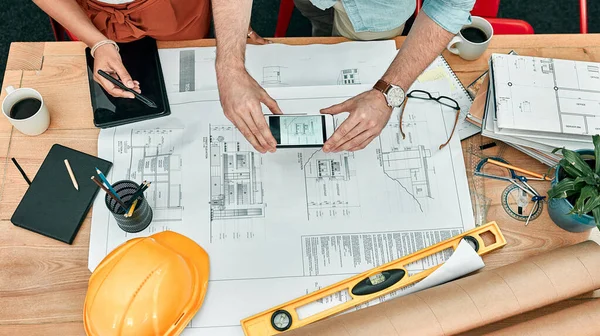 Its so simple to save and share their ideas. Closeup shot of two unrecognisable architects using a cellphone to take photos of blueprints in an office