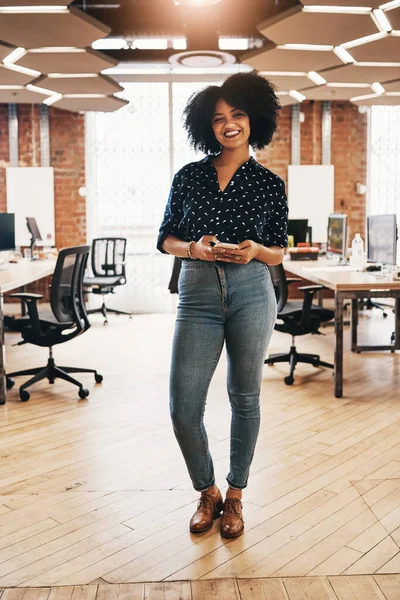 Im always online and chatting with clients at work. Full length portrait of a cheerful young female designer holding a cellphone while standing inside of her office
