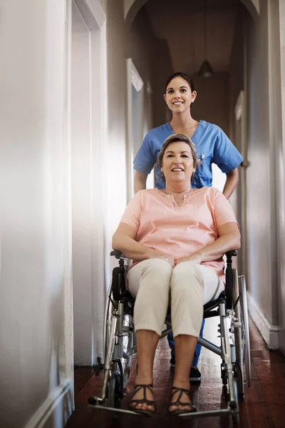 彼女は周りに行くことが容易になります 老人ホームの車椅子で高齢者を押す魅力的な若い女性介護者の完全な長さのショット — ストック写真