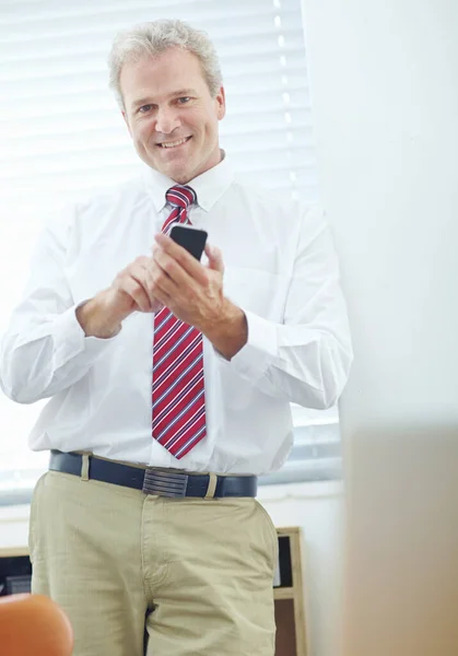 Mobile business with a positive result. Attractive businessman leaning against the office wall while looking at the camera and holding his mobile phone