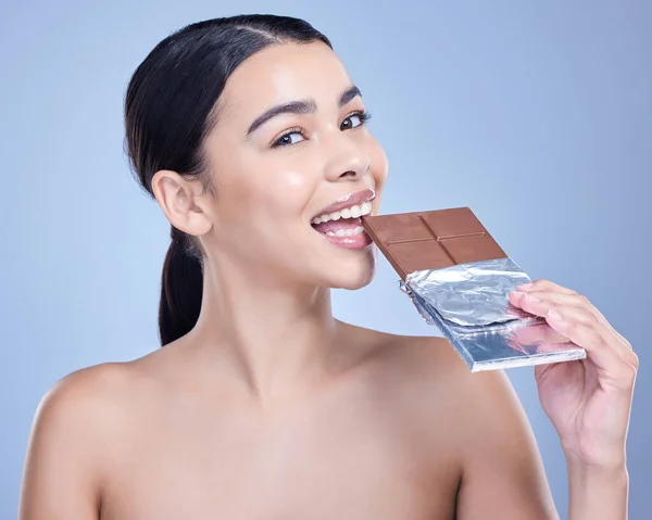 Studio portrait of a beautiful mixed race woman holding a slab of chocolate. Hispanic model snacking on dessert against a blue copyspace background.