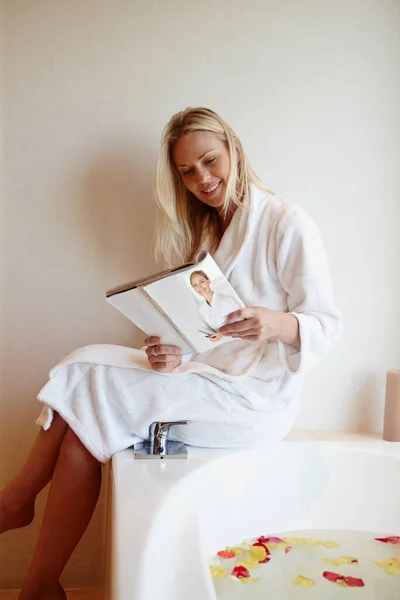 私の個人的なスパ 花びらでいっぱいの豪華な風呂の端に座っている間に雑誌を読んでいる美しい若い女性 — ストック写真