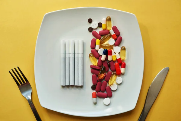 Dieting takes a dangerous turn. Studio shot of medication and cigarettes served on a plate with a fork and knife against a yellow background