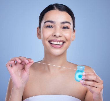 Studio portrait of a smiling mixed race young woman with glowing skin posing against blue copyspace background while flossing her teeth for fresh breath. Hispanic model using floss to prevent a cavit.