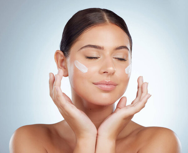 Beautiful woman applying face cream while posing with copyspace. Caucasian model isolated against grey studio background with product on cheek. Moisturise and sunscreen for healthy skincare routine.