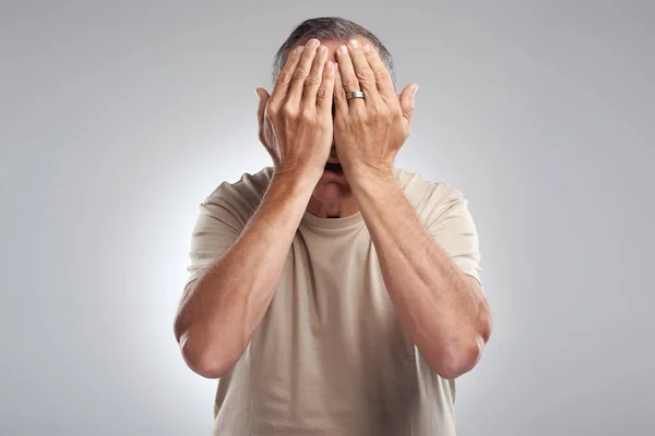 See no evil. a mature man standing against a grey background in the studio and covering his face with his hands