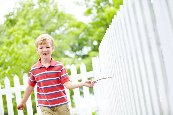 惊慌失措地走过警戒线 一个可爱的小男孩在自家后院的白色篱笆上跑着一根棍子 — 图库照片