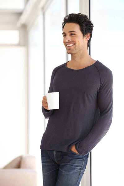 Happy, smile and man with a cup of coffee standing by the window in a vacation house. Calm, relax and attractive male person drinking a latte in a mug in the living room while thinking or in a dream