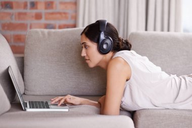 Kadın, dizüstü bilgisayar ve kulaklıkla evde müzik dinlemek ya da web videosu aramak için kanepede uzanmak. Oturma odasındaki kanepede rahatlamak için kız, bilgisayar ya da film, podcast ya da ses kaydı.