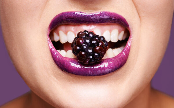 Woman lips, purple lipstick with berry and makeup, shine and creativity with beauty isolated on studio background. Closeup of fruit between female model teeth, cosmetic product and cosmetology.
