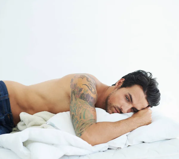 Thinking, relax and topless with a sexy man on a bed, lying in studio on a white background. Tattoo, idea and shirtless with a handsome young male model posing in a bedroom for sensuality or desire.