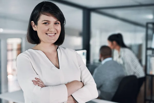 stock image Theres nothing my team cant achieve. an attractive young businesswoman standing with her arms folded while colleagues works behind her