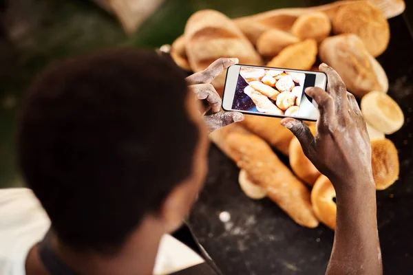 People like to see what youre up to. a male baker taking a picture on his cellphone of a selection of freshly baked bread