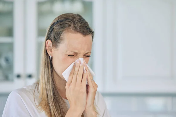 Sick, tissue and woman blowing her nose in the kitchen for a cold, flu or sneeze at her home. Illness, virus and young female person sneezing with allergies, hay fever or covid in her apartment