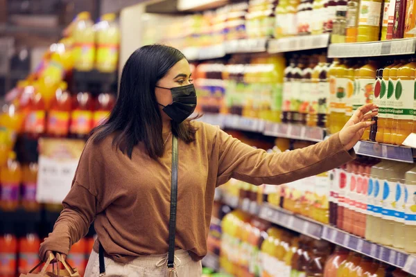 Grocery shopping, woman and juice at a shop, market and store for groceries sale with mask. Health, virus safety and female person with orange and healthy drink purchase in a supermarket at shelf.
