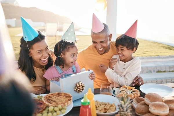 stock image Happy birthday, love and family outdoors for celebration, party and present, laugh and excited. Parents, children and gift for girl child at special event, smile and celebrating with food outside.