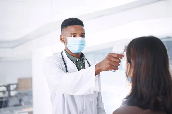 这只需要一秒钟 一位戴口罩的年轻医生 她和一位病人站在诊所里量体温 — 图库照片