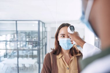 Umarım ateşiniz falan yoktur. Maske takan genç bir kadın ve klinikteki doktoru ateşini ölçtürüyor.