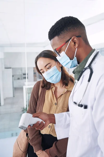 你今天跑得有点高了 一位戴口罩的年轻医生 她和一位病人站在诊所里量体温 — 图库照片
