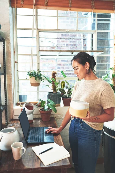 stock image Time to put this pot up for sale. an attractive young business owner standing and holding a clay pot while using her laptop