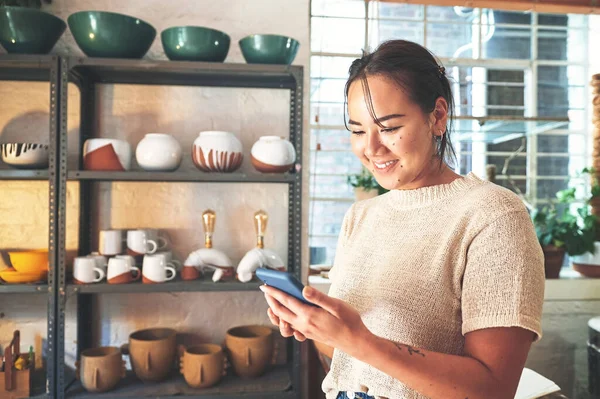 Staying in touch with clients via text. an attractive young business owner standing alone in her pottery studio and using her cellphone