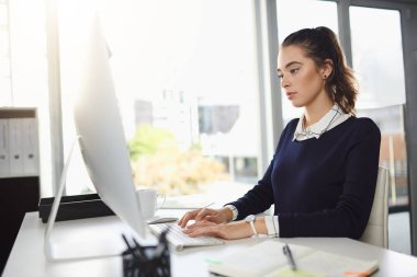 Bugün yapmam gereken işe odaklanmış durumdayım. Çekici genç bir iş kadını masasında oturuyor ve bilgisayarını modern bir ofiste kullanıyor.