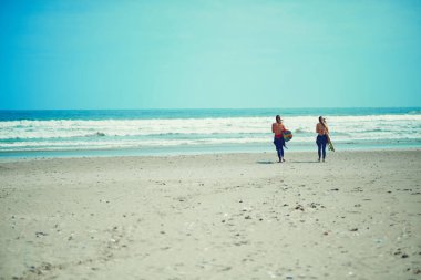 Okyanus beklemez. Sahilde sörf tahtasıyla yürüyen genç bir çift.