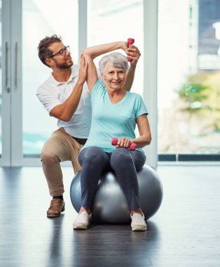 Kendini spora adadı. İyileşme sürecinde erkek bir fizyoterapistle çalışan yaşlı bir kadının tam boy portresi.