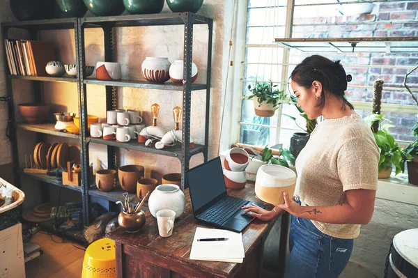 stock image How much stock do I have of this. an attractive young business owner standing and holding a clay pot while using her laptop