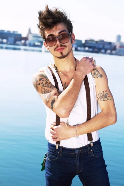 Sunglasses, portrait and serious man by sea with stylish tattoo, body art and fashion. Punk, ocean and trendy male person standing outdoor in Spain with cool clothes, attitude and aesthetic mockup