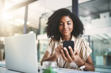 Mobil uygulamalar bir şeyler yapmasına yardımcı oluyor. Modern bir ofisteki masasında cep telefonu ve laptop kullanan genç bir iş kadını.