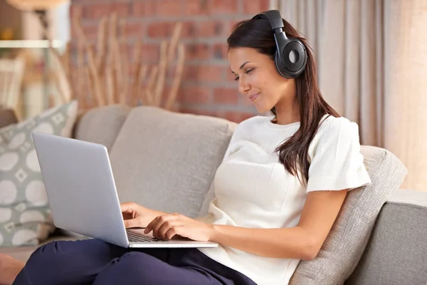 stock image Home, headphones and a woman typing on a laptop and listening to music or audio while streaming online. Happy female person relax on sofa to listen to radio or watch a movie with internet connection.