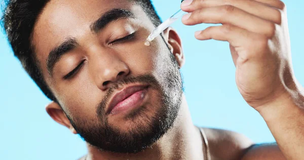 Skincare, health and man with face serum in a studio with a beauty, grooming and face routine. Wellness, cosmetic and male model with a facial oil pipette for a skin treatment by a blue background