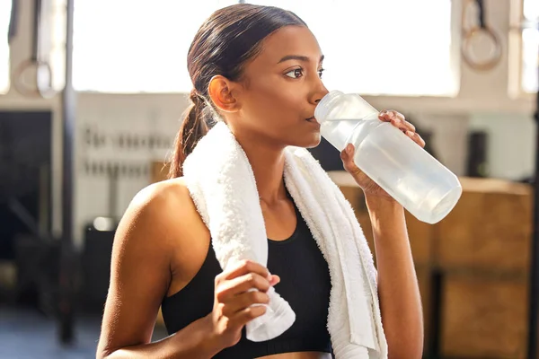 Tired, fitness or Indian woman drinking water at gym in training, workout or exercise to hydrate her body. Fatigue, wellness or thirsty girl with bottle for healthy liquid hydration on resting break.