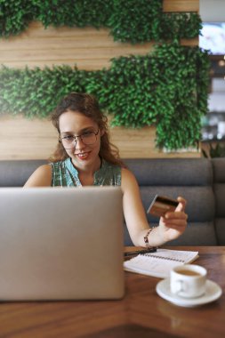 Banka kuyruklarında daha az zaman, işini büyütmek için daha çok zaman. Bir kafede çalışırken bilgisayar ve kredi kartı kullanan genç bir kadın.