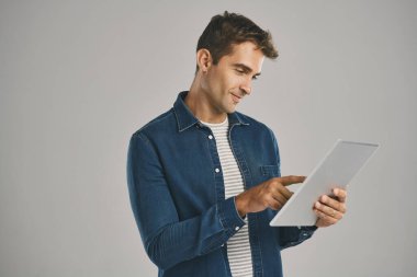 İnternetteki herkesle bağlantı kuruyorum. Gri arkaplana karşı dijital tablet kullanan genç bir adamın stüdyo fotoğrafı.