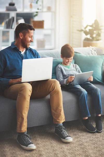 stock image They are both working from home. a cheerful little boy and his father using a laptop and a digital tablet while being seated on a sofa at home during the day