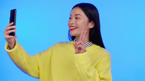 亚洲女性 自拍与和平标志为社交媒体形象图片的蓝色背景 在智能手机上聊天 视频通话和亲吻的移动应用程序上快乐的模特 — 图库视频影像