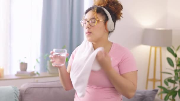 毛巾和女人在家里客厅里锻炼后喝水 水合和疲惫的年轻女性在家里做完音乐后休息时汗流浃背 — 图库视频影像