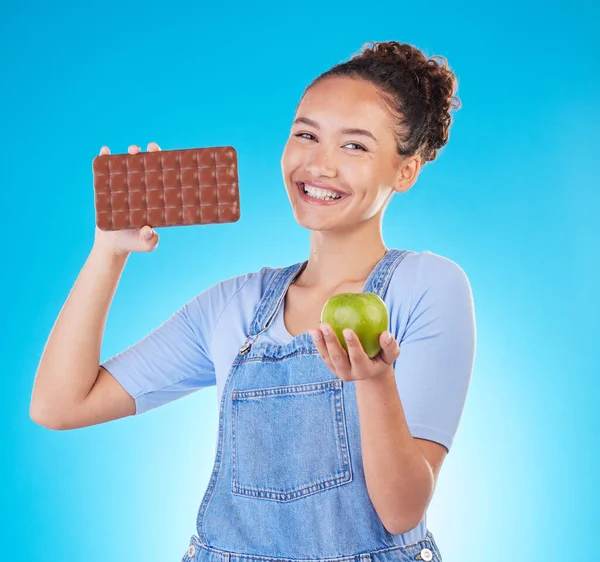Apple, chocolate and woman for healthy food choice or offer isolated on studio, blue background for food, sugar and diet. Dessert, green fruit and happy young person for detox or lose weight decision.