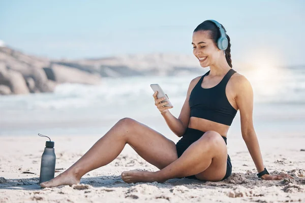 stock image Fitness, break and happy woman relax at beach with phone, music and headphones after training. Exercise, resting and lady health influencer posting to social media, blog or podcast after ocean run.