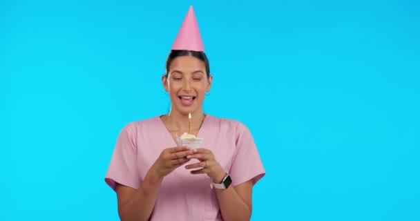 誕生日 パーティー お祝いのために青の背景に隔離されたスタジオで看護婦の女性と願います 幸せな若い女性医療専門家カップケーキの上にろうそくを吹き消す — ストック動画