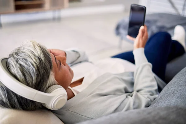 Senior woman, headphones and phone on sofa, relax and music in home living room with search, choice and listening. Elderly lady, smartphone and audio streaming app for radio, podcast and lounge couch.