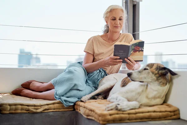 Next to dogs, books are my best friends. Shot of a mature woman reading a book on her balcony with her dog next to her at home