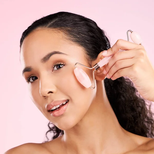 Face roller, natural beauty and woman with skin glow, health or wellness cosmetics. Headshot of a young aesthetic female model with dermatology and facial massage results on a pink background.