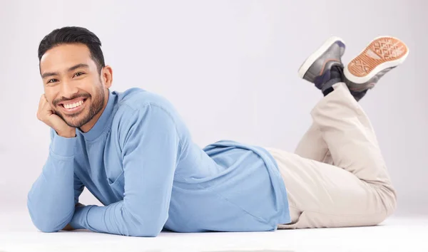 Portrait, happy or Asian man lying on floor isolated on a white background in studio to relax. Friendly smile, calm person or proud male model resting with confidence, fashion or style on the ground.