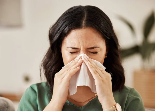Blowing nose, tissue and woman in a living room with flu, cold and hay fever, crisis or viral infection in her home. Sneezing, allergies and person with virus or coughing illness in a lounge in house.